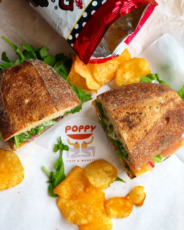 Two sandwich halves on paper with potato chips, an open chip bag and a Poppy sticker, seen from above