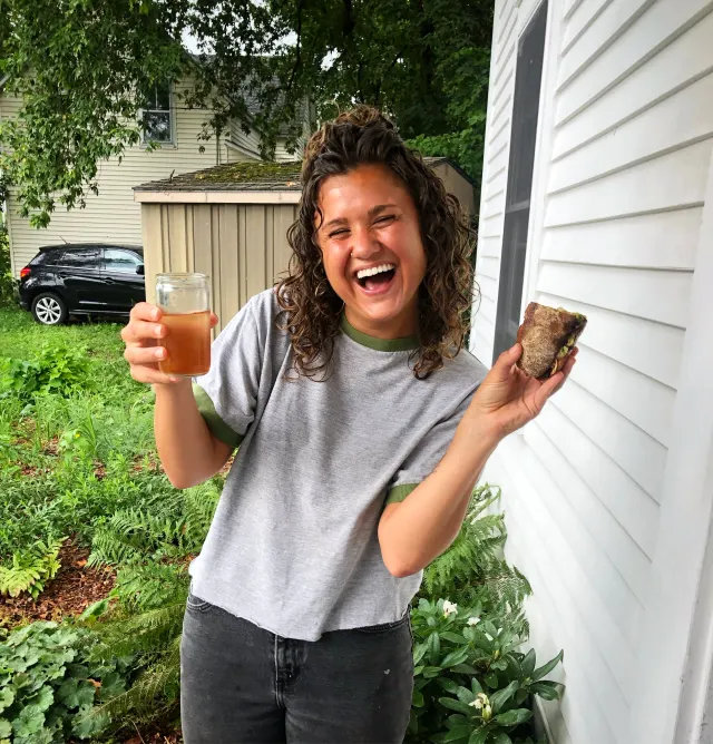 A person laughing in a garden beside a white house, holding up a glass