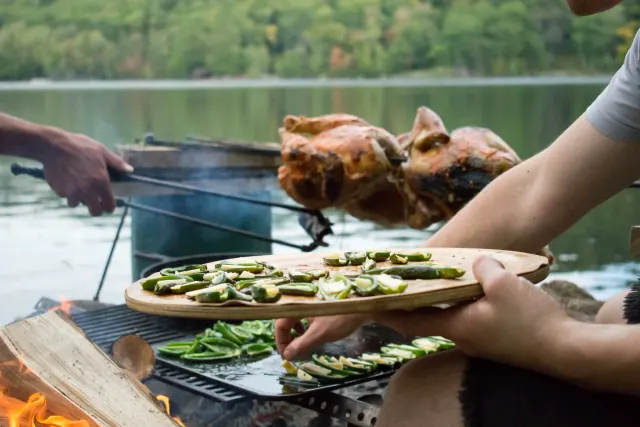 Hands at an open fire by a lake, peppers on a wooden board and a roast on the grill
