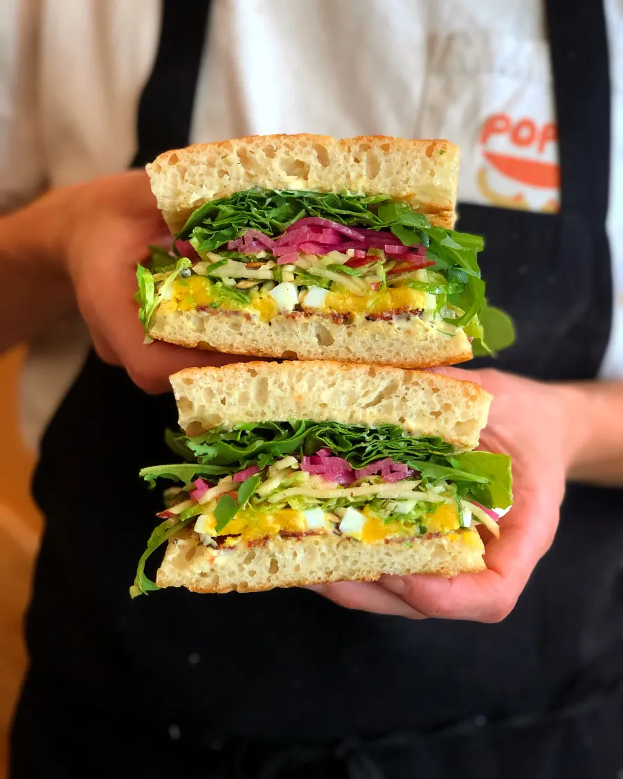 Two generous sandwich halves held toward the camera, with greens and pickled onions between focaccia