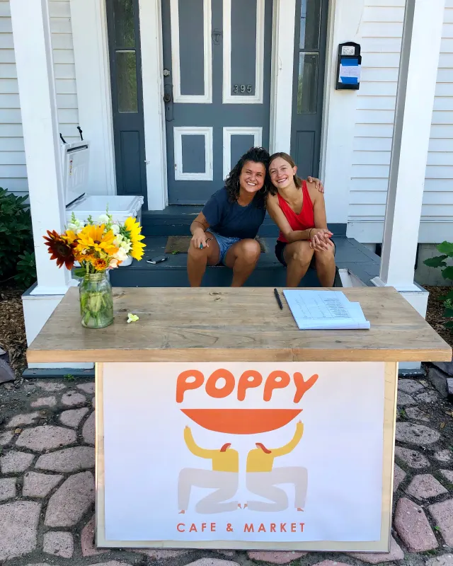 Two people sitting on porch steps behind a wooden table with a Poppy Cafe & Market sign and a jar of sunflowers