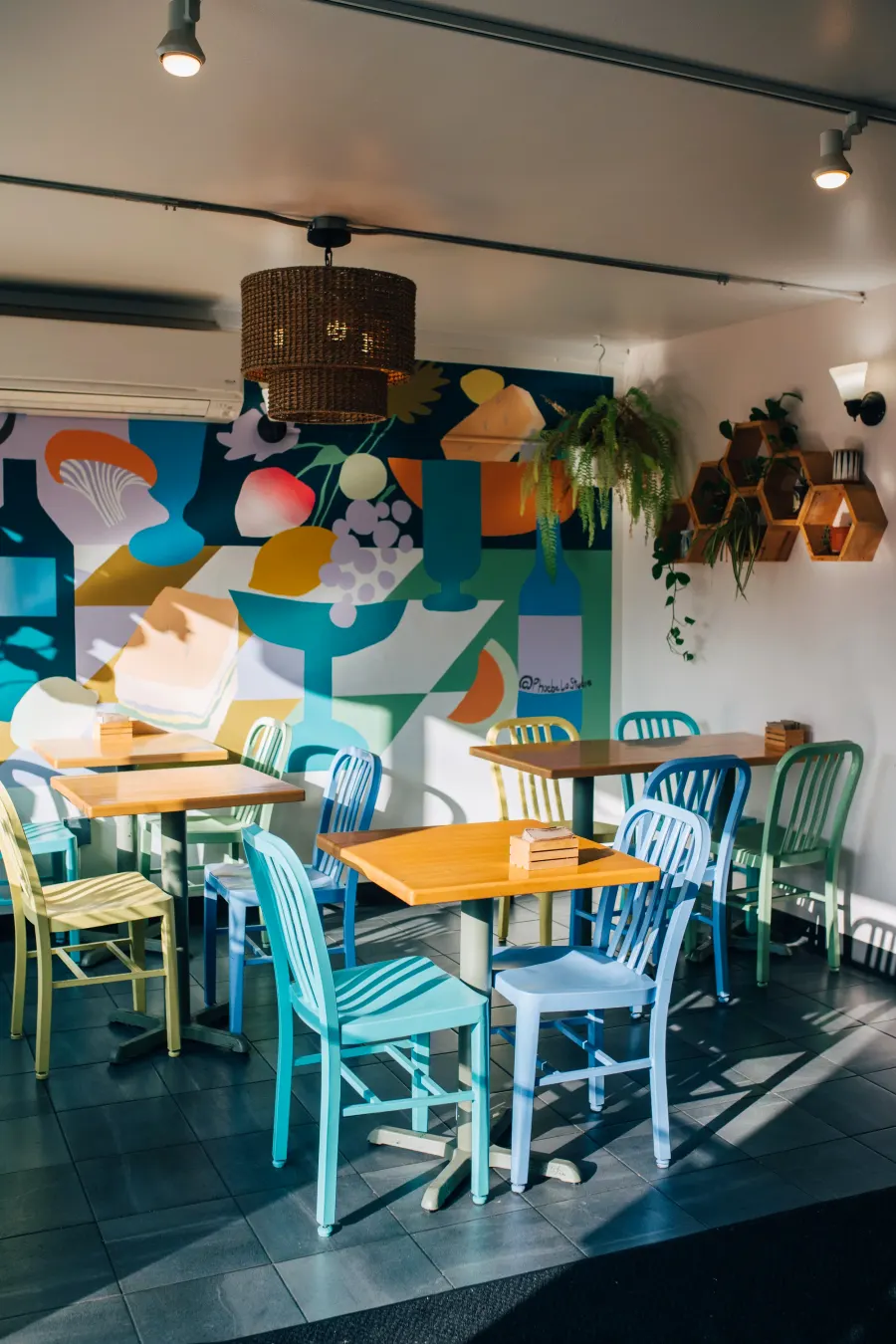A sunlit dining room with painted chairs, wooden tables, a woven lamp and a colorful mural