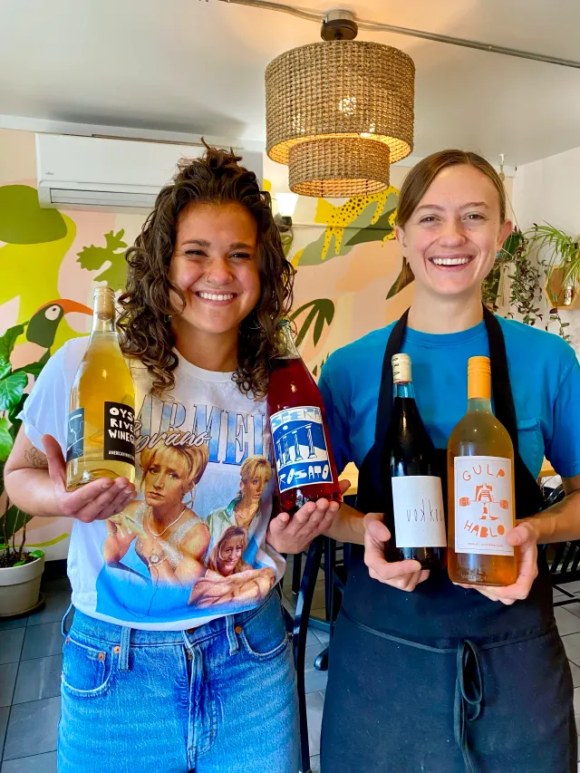 Two people smiling and holding up bottles in front of a painted wall