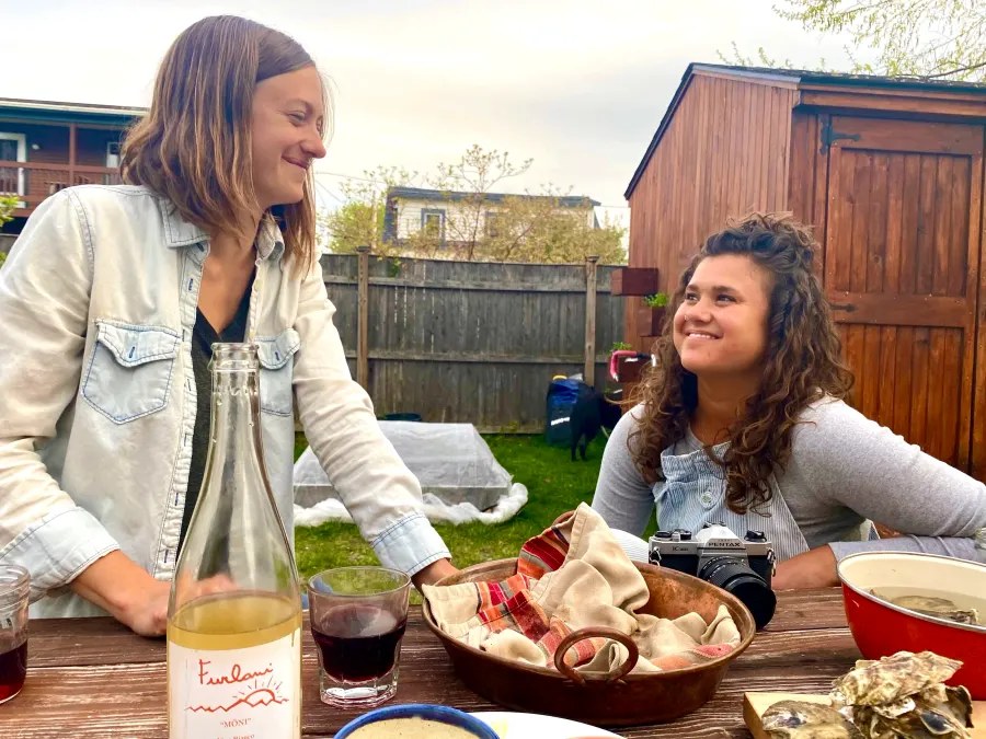 Two people at an outdoor table with a wine bottle, glasses, a woven bowl, a camera and a red pot