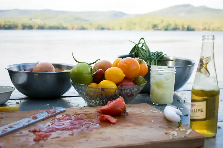 Bowls of tomatoes, citrus and green beans beside a cutting board, a lake and hills behind
