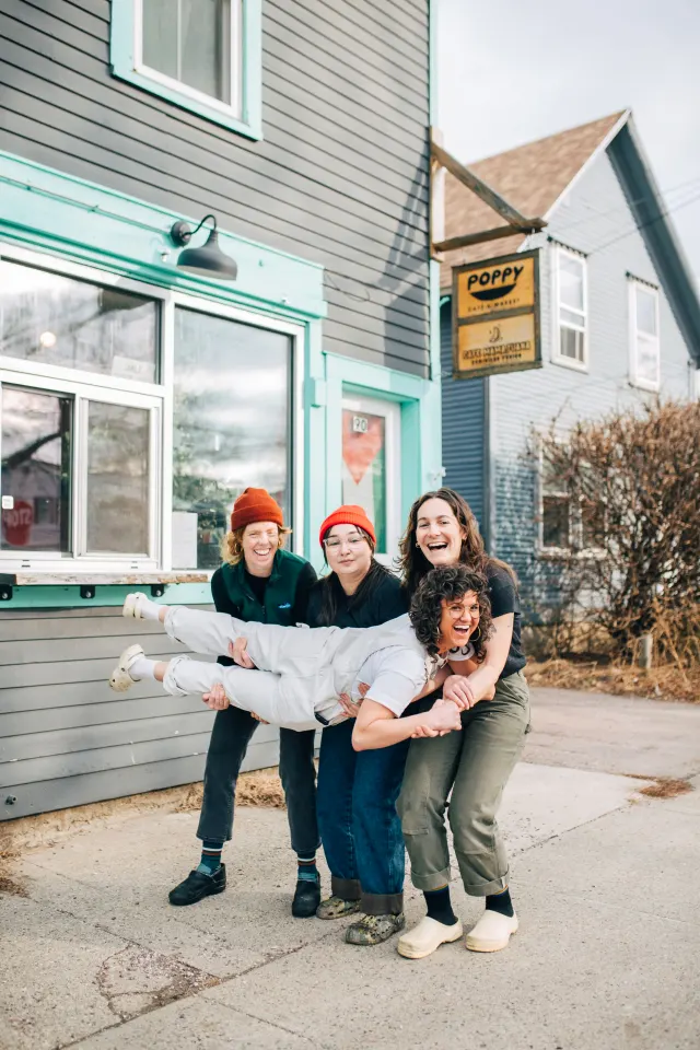 Three smiling people hold a fourth up sideways on the sidewalk under a hanging Poppy sign