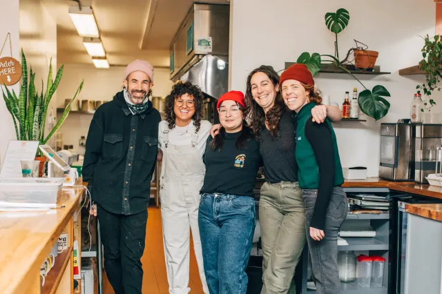 Five people standing arm in arm in a kitchen with plants on the shelves
