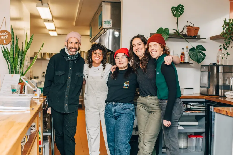 Five people standing arm in arm in a kitchen, plants on the shelves behind them